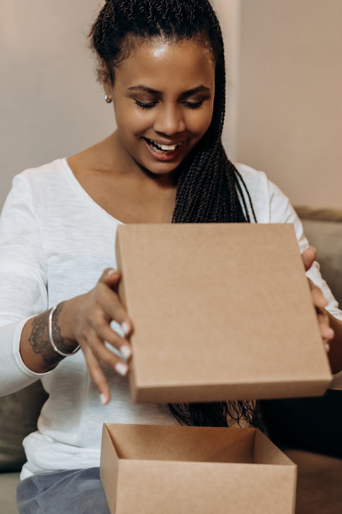 Smiling woman eagerly opens a cardboard gift box indoors, revealing excitement.